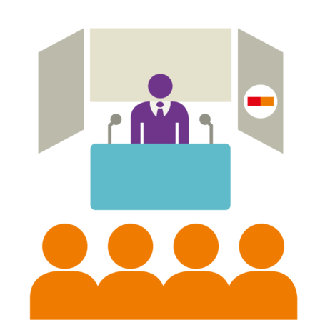 Vier Personen schauen auf eine Person, die an einem Redepult steht.